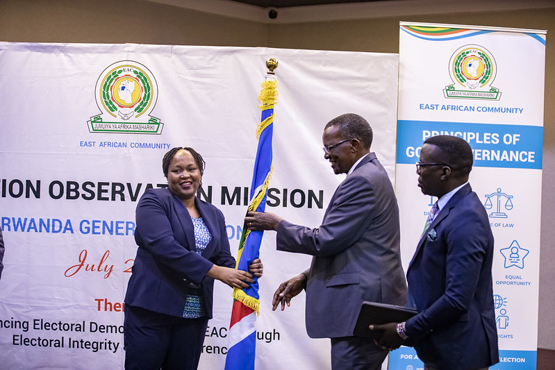 The EAC Secretary General, H.E. Veronica Nduva, presents the EAC Flag to signify the handing over to the Head of the EAC Election Observation Mission to Rwanda, Chief Justice (Emeritus) David Kenani Maraga, during the launch of the mission in Kigali, Rwanda. Looking on (extreme right) is the Deputy Head of Mission, Hon. Siranda Gerald Blacks, of the East African Legislative Assembly.